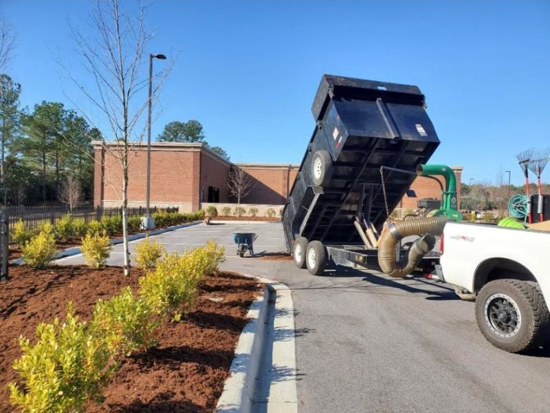 Landscaping crew spreading mulch along shrubs beside a commercial building with a dump trailer and truck